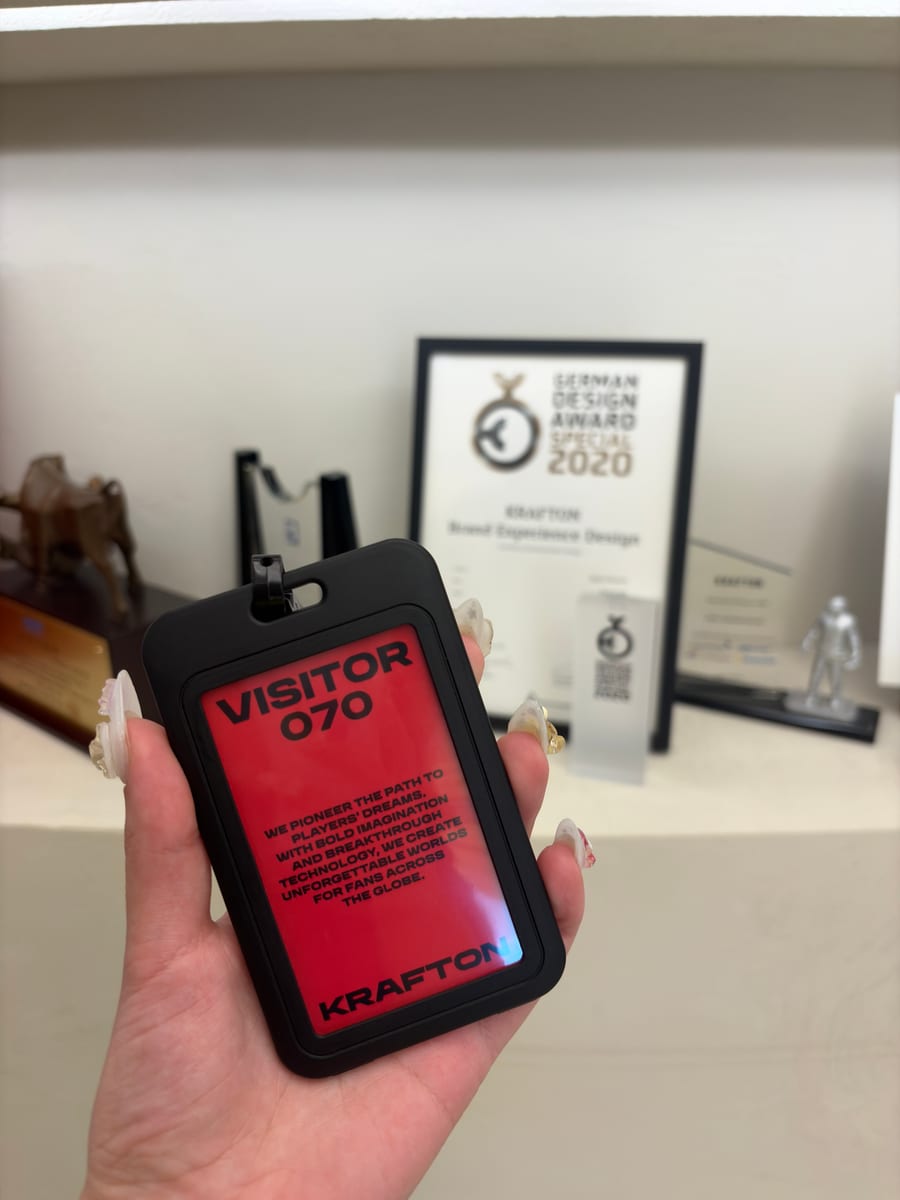 A hand holding a red KRAFTON visitor badge in front of the company's design awards